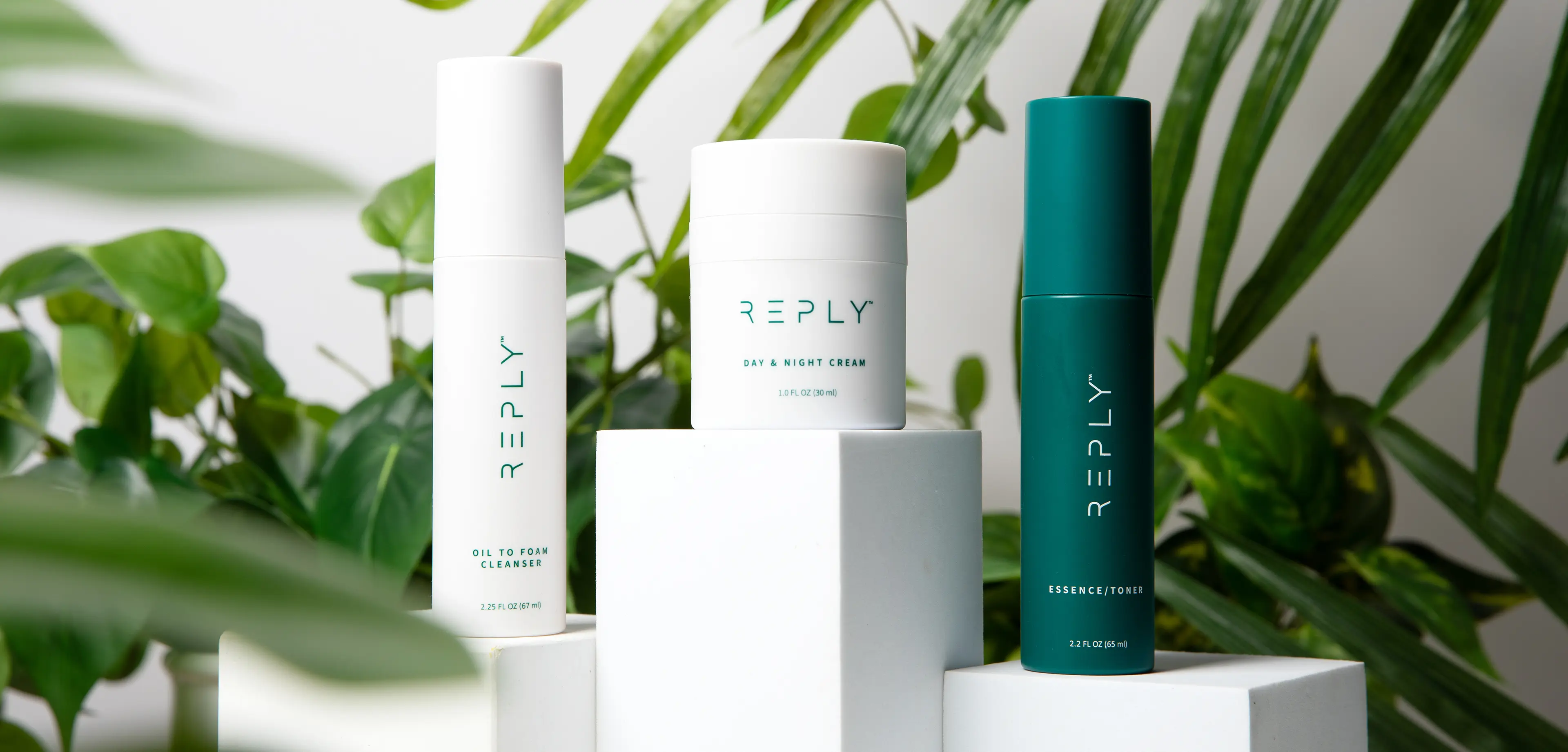 A picture of containers of REPLY Oil To Foam Cleanser, REPLY Day and Night Cream and REPLY Essence/Toner arranged on white boxes of different heights in front of a background of green house plants.  At the bottom is a button labeled 'Shop Now' 