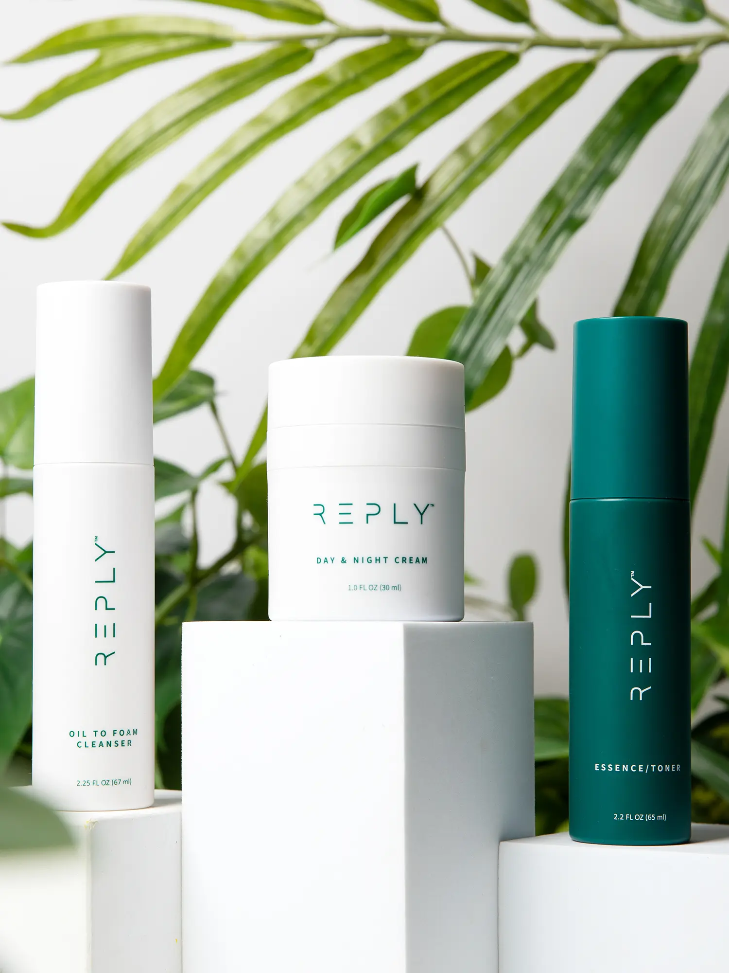 A picture of containers of REPLY Oil To Foam Cleanser, REPLY Day and Night Cream and REPLY Essence/Toner arranged on white boxes of different heights in front of a background of green house plants.  At the bottom is a button labeled 'Shop Now' 